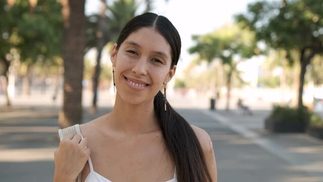young latin american woman with long dark hair looking at camera and smiling. She is in a green park with palm trees in Barcelona during a sunny summer day