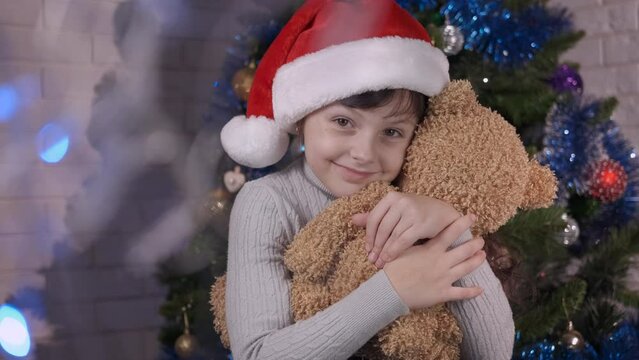 Embrace teddy at Christmas. A happy joyful child in Santa hat embrace her teddy bear in the room by the Christmas tree.