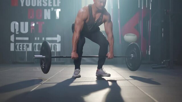 Focus of young Asian muscular fitness man's hands and legs while doing a barbell deadlift by bend down to grasp the barbell on the floor and lift it up at the gym with light shines through the window.