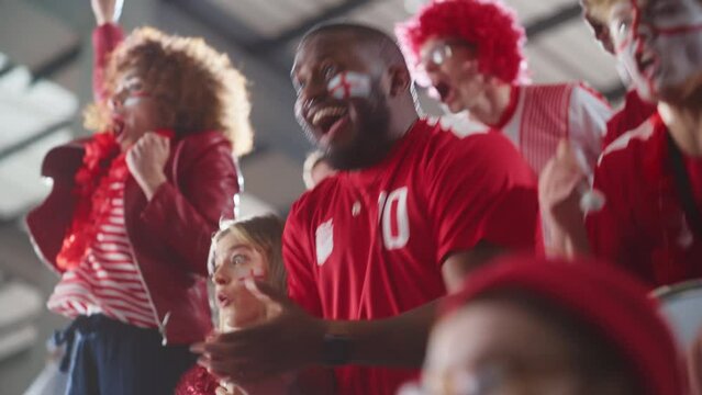 Sport Stadium Big Event: Handsome Expressive Black Man with Painted face Jumping Up and Cheering Goal Score. Crowd of Fans Shout for Red Soccer Team to Win. People Celebrate Championship Victory