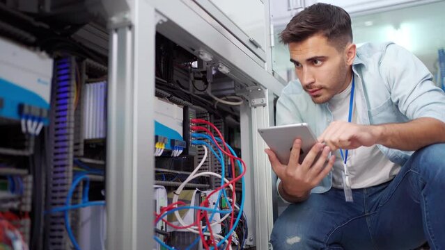Young Caucasian male electrical engineer in blue shirt squatting touching wire cables and using a digital tablet checking the electrical system of the industrial machine in an industrial factory.