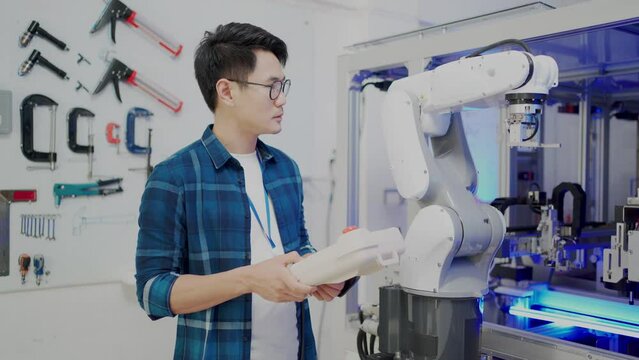 Young Asian male industrial engineer in blue plaid shirt and glasses standing testing the work of an automation robotic arm machine with a robot controller in his hands in an industrial factory.