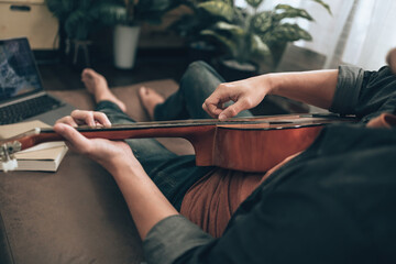 young man relax and playing guitar while sitting on sofa bed in living room at home. Music create melody song, lyrics on laptop and practice concept.