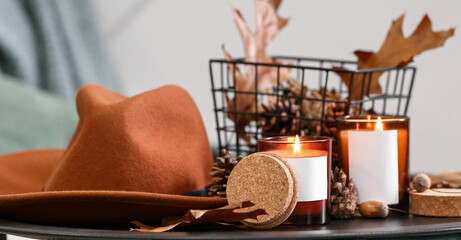 Burning candles in holders, felt hat and autumn decor on table in room