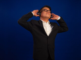 Portrait of smart middle-aged asian confident successful businessman wearing suit listening music with wireless headphones isolated on blue background in studio, looking happy leadership