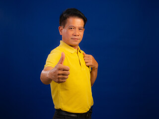 Portrait of smiling middle-aged asian man in casual yellow polo shirt showing thumb up isolated on blue background in studio, looking happiness and attractive professional leadership