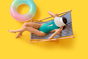 Beautiful young woman in swimsuit relaxing on deck chair against yellow background, top view