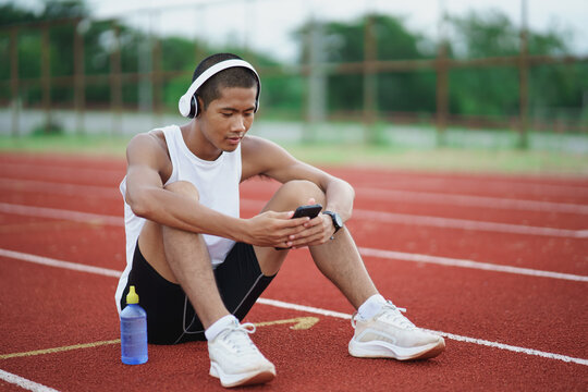Asian man sportsman fit body wearing white headphone and using mobile phone to listen music while resting or before jogging, exercising at lane stadium. Sport healthy running concept.