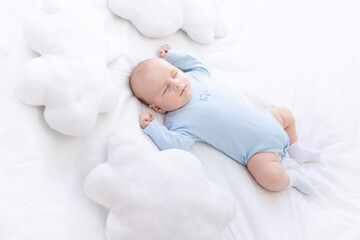 baby boy sleeps on the bed lying on his back in blue pajamas with his hands up among the pillows of...