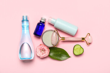 Naklejka premium Facial massage tool with cosmetic products, cucumber slice and flower on pink background