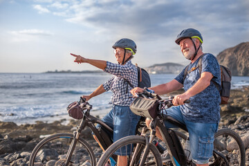 Senior caucasian couple riding off road on the pebble beach with bicycles at sunset looking away...