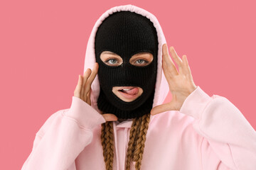 Young woman in balaclava with hands near face showing tongue on pink background