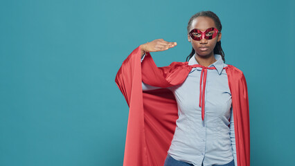 Portrait of woman superhero doing military salute to show motivation and respect. Wearing hero costume with red cape and mask, feeling confident and powerful over blue background in studio.