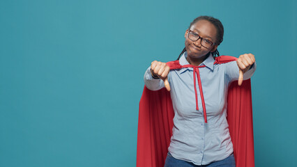 Portrait of woman giving thumbs down and wearing red cape to be cartoon character superhero with...