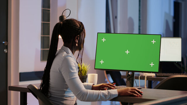 African american woman looking at computer with greenscreen in office late at night. Analyzing isolated chroma key template with blank mockup background and copyspace monitor display.