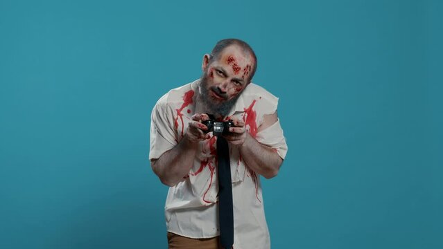 Spooky bizarre looking zombie with modern gaming controller playing games on console. Sinister mindless brain-eating monster playing with electronic wireless gamepad on blue background.