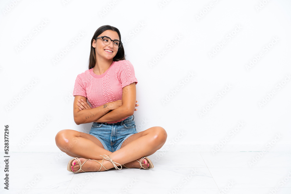 Wall mural young caucasian woman sitting on the floor isolated on white background with arms crossed and happy