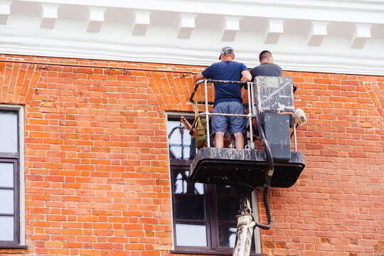 Repair Of The Facade Of A Brick House. Two Workers On An Aerial Platform Inspect The Wall And Identify Defects After Major Repairs. Unrecognizable Person