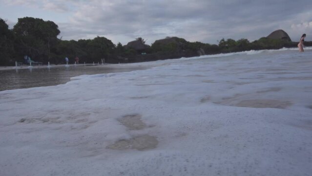 View of a woman standing in the sea waves reaching the sand of the shore under the cloudy sky