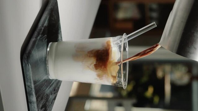 Vertical Video home barista making iced latte pouring espresso shot into glass drinking cup containing milk with glass straw in slow motion close up