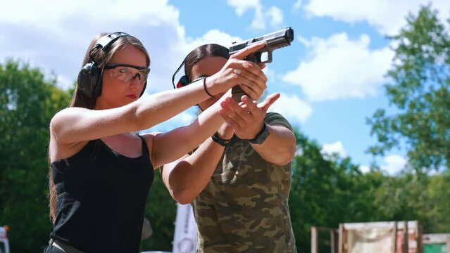 A day in a shooting range. Confident caucasian young adult girl aiming at target with a black gun. Protective eyeglasses and headphones. Private shooting lesson. High quality 4k footage