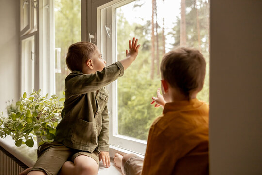 Children sitting on windowsill and waiting for someone comming. Two brothers, friends. Cute preschool kids alone at home. Boys are waiting for their mother or father. Loneliness. Busy parents.