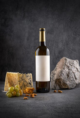 Naklejka premium Glass and bottle of white wine with grapes on dark background. White wine with cheese and fruits. Wine bottle mockup with blank labels to place your design. Space for text