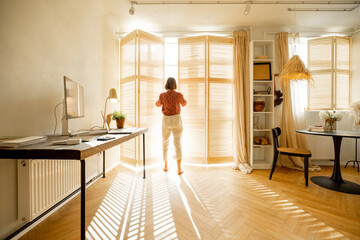 Woman opens window blinds letting the sun inside the room, spending good morning in sunny and cozy apartment in beige tones. Interior view - Powered by Adobe