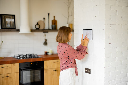 Woman controls room temperature on a digital panel mounted on the wall in kitchen. Concept of smart home and new technologies for home comfort