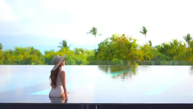 Back View of Sexy Woman in Swimsuit and Hat in Infinity Pool With Beautiful View of Tropical Island and Ocean Horizon