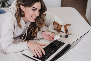 Young woman with credit card doing online shopping through laptop lying by dog at home