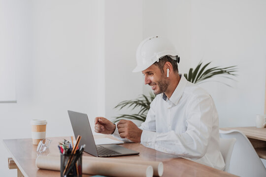 Smiling engineer wearing hardhat having video call on laptop at desk in office