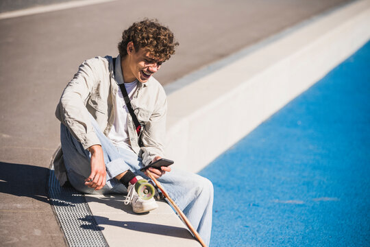 Happy man with skateboard using smart phone at edge of track and field