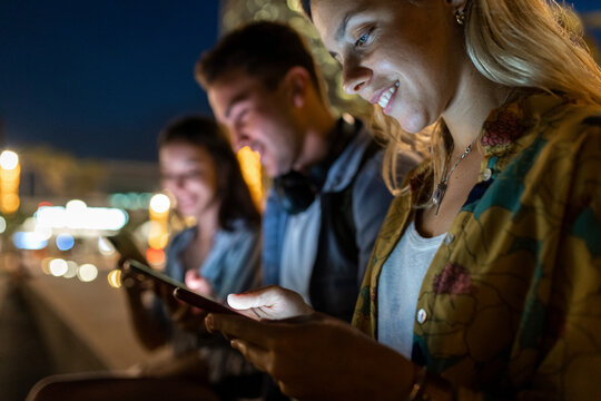 Smiling young woman using mobile phone sitting by friends at night