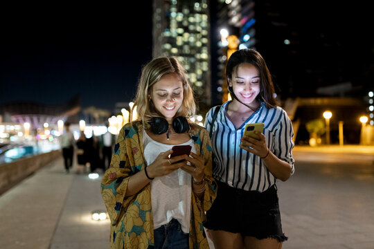 Smiling young women using mobile phones in city at night