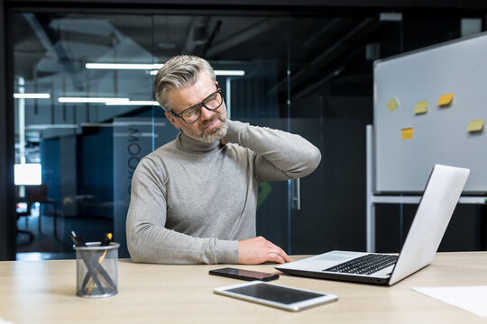 Senior mature businessman working inside office building, gray haired man sick and tired massaging neck with hand, boss having severe pain in neck and back.