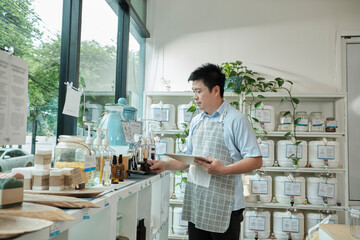 Asian male shopkeeper checks stock of natural organic products at window display in refill store, zero-waste and plastic-free grocery, eco environment-friendly, sustainable lifestyles, reusable shop. - obrazy, fototapety, plakaty
