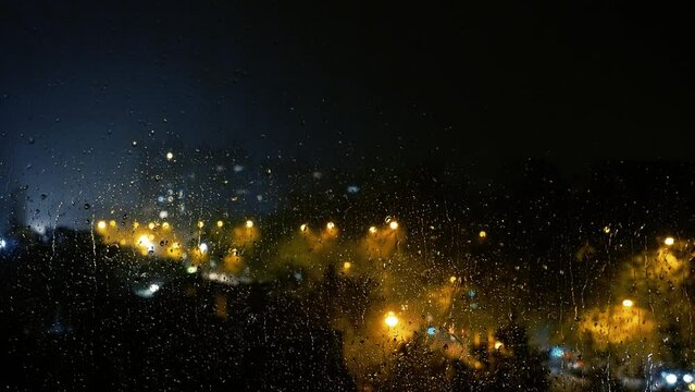 Raindrops On Window Against Night City Street During Thunderstorm. View of Blurred City Night Illumination Through The Window With Raindrops. City Lights And Traffic On A Rainy Night. Melancholy Rain.