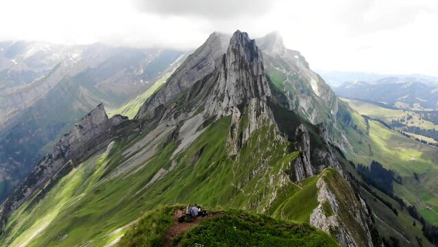 Aerial flyover over the two hikers enjoying the view of  Schafler ridge in Appenzell, Switzerland with cliffs and mountain peaks in view