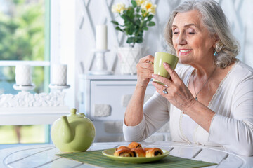 Obraz na płótnie Canvas Beautiful smiling senior woman drinking tea while sitting at kitchen table