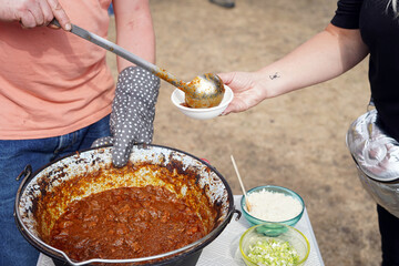 Serving the stew that was prepared in the cauldron. It is poured into a plastic bowl with a metal ladle.