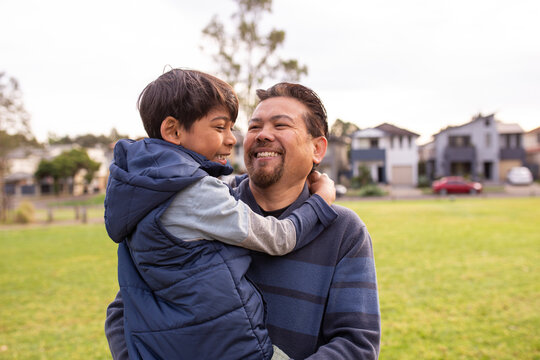 smiling middle aged man wearing blue sweatshirt carrying a smiling boy in his arms on a big lawn