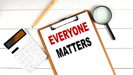 EVERYONE MATTERS words on clipboard, with calculator, magnifier and pencil on the white wooden background