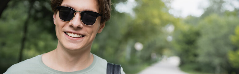 young and happy tourist in sunglasses looking at camera on blurred background, banner.