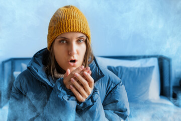 A woman in a warm jacket and hat sits on the bed at home and tries to keep warm. Home heating...