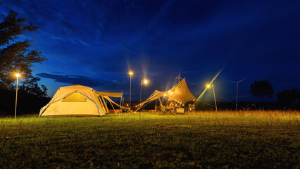 Outdoor camping tent with tarp or flysheet on grass courtyard and warm night light under dark blue...