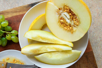 sliced melon with seeds on a white plate, a kitchen knife and green grapes on a wooden board, the concept of fresh fruit and healthy eating