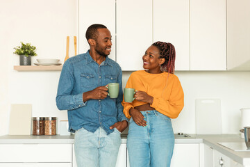 Fototapeta premium Happy african american spouses drinking morning coffee and talking, standing in kitchen interior, copy space