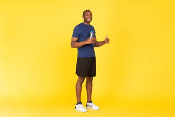 Fototapeta na wymiar African Male Gesturing Thumbs Up Holding Fitness Bottle, Yellow Background