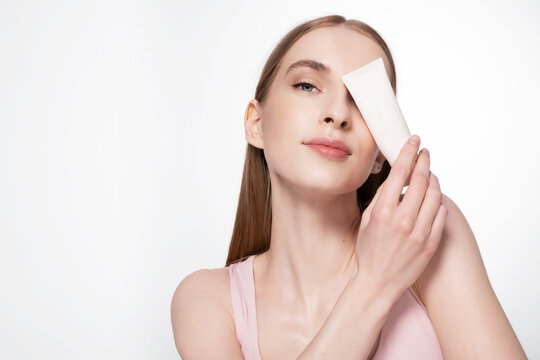 Girl with perfect light skin holding mockup cosmetics tube of skincare cream or make up foundation on white studio background with copy space. Natural woman beauty. Cosmetic products advertising